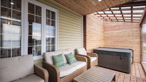 A covered outdoor seating area with wicker furniture, a hot tub, and a view through large windows.