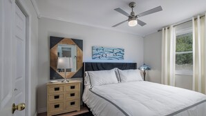 A bedroom with a bed, a ceiling fan, a nightstand, a lamp, a dresser, and a painting.