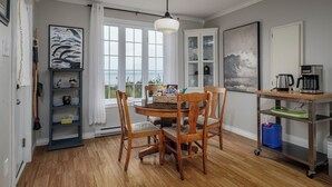 A dining area with a round table, wooden chairs, a chandelier, a view through large windows, a cabinet with glass doors, and a coffee station.