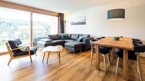 A modern living room with a wooden floor, a sectional sofa, a dining table with chairs, and a large window offering a view of the outdoors. - Nice apartment for 6 guests with WIFI, TV, patio and panoramic view (Waldhaus)