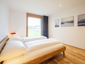 Una cama doble con estructura de madera, un ventanal con vistas a un paisaje verde y tres cuadros colgados en la pared.
