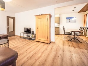 Ein Wohnzimmer mit Holzschrank, Fernseher auf einem Schrank, Esstisch mit Stühlen und einer Küchenzeile im Hintergrund.