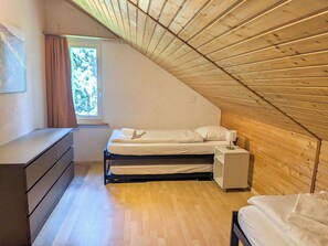 Quarto com teto inclinado, cama, mesa de cabeceira, cômoda e janela com vista para árvores.