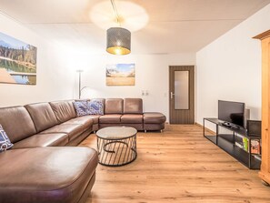 Ein modernes Wohnzimmer mit einem ledernen Ecksofa, einem runden Couchtisch, einem Fernseher auf einem schwarzen Schrank und einem Bücherregal.