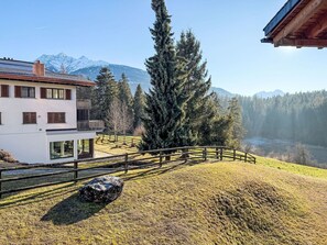 Ein modernes, zweistöckiges Haus mit Balkon, umgeben von einem Holzzaun, einem großen immergrünen Baum und Blick auf schneebedeckte Berge.