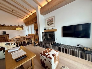 Ein gemütliches Wohnzimmer mit Kamin, Holzmöbeln, einem Fernseher und einem Hocker aus Rindsleder.