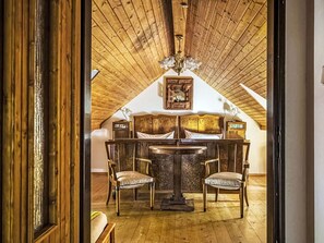 An attic bedroom with a wooden ceiling, a chandelier, a bed with headboard, two chairs, and a table.