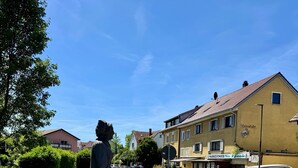 Eine Statue auf einem Platz mit einem Brunnen, umgeben von Gebäuden und blauem Himmel.