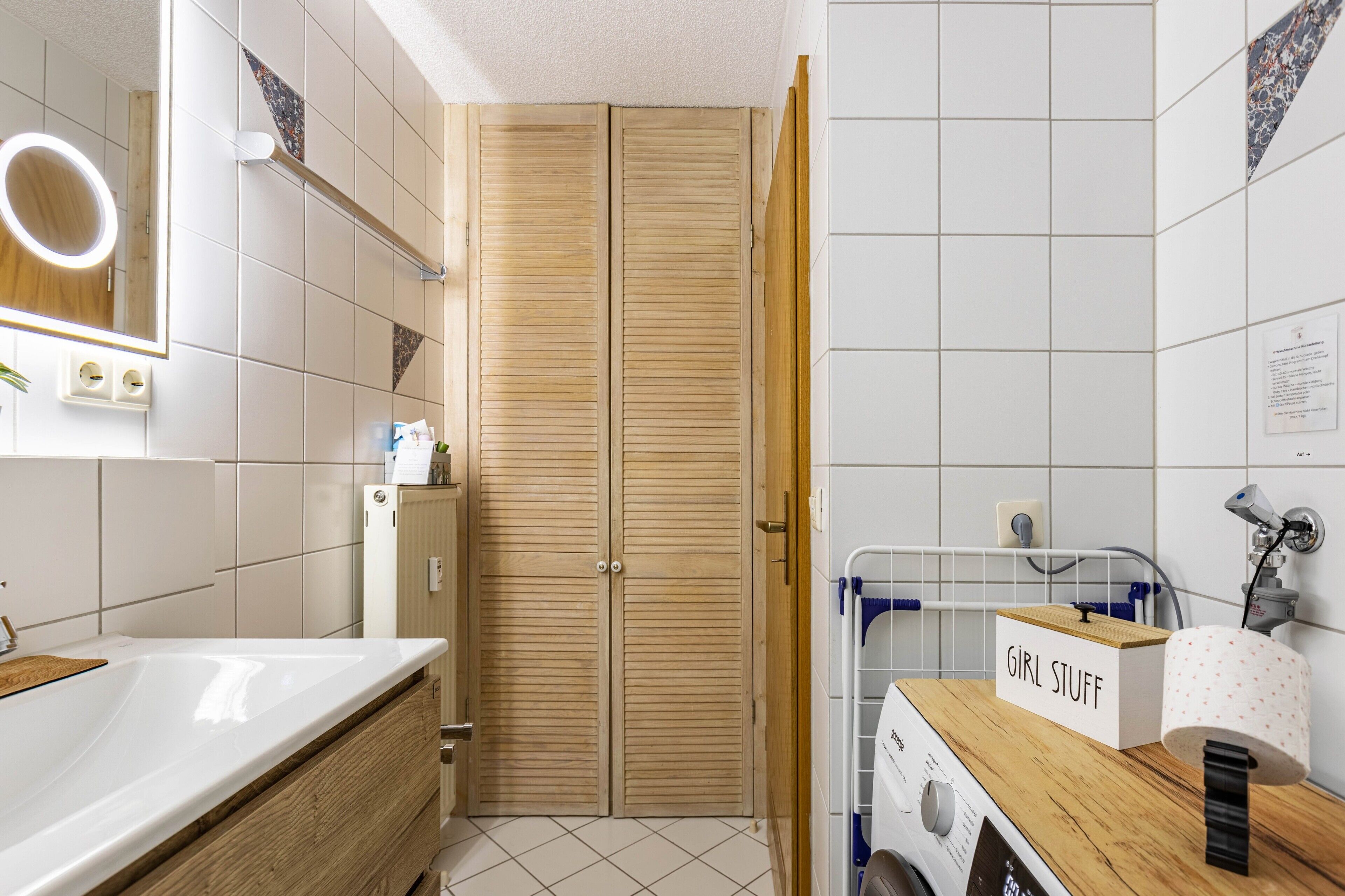 A bathroom with a wooden vanity, a sink, a mirror with a light, a towel rack, and a laundry machine.