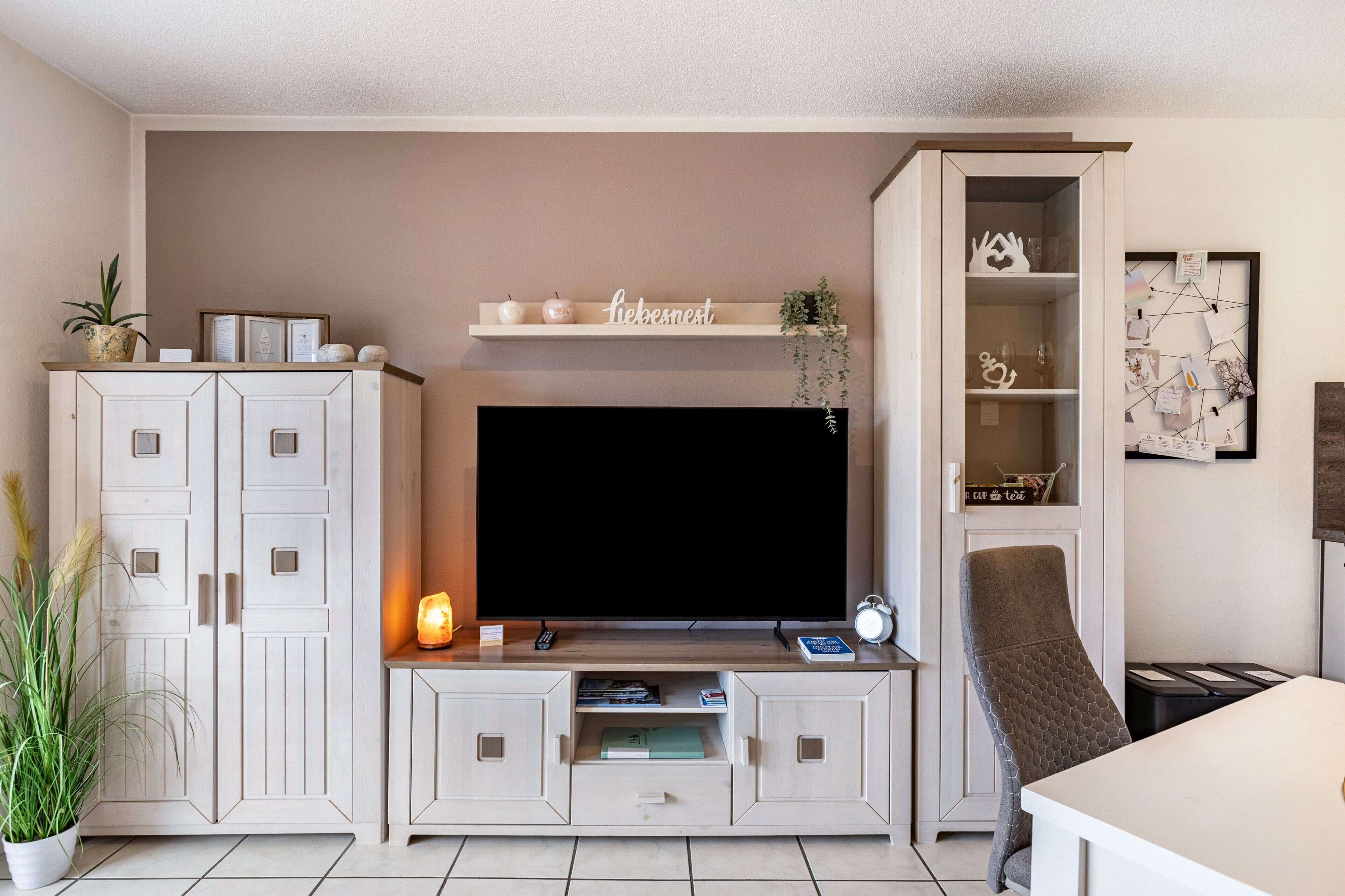 Ein Wohnzimmer mit Fernseher, Schränken, einem Sessel und einem Regal mit Dekoration.
