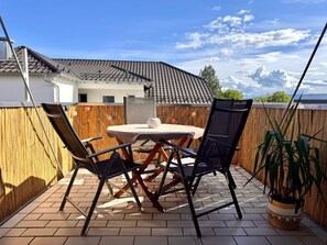 Eine Terrasse mit rundem Tisch und Stühlen, einem Blumentopf und Blick auf ein Haus mit Ziegeldach.