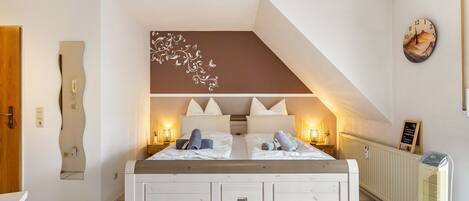 Una camera da letto con un letto bianco, due abat-jour, un elemento decorativo a parete e una sveglia appesa al muro.