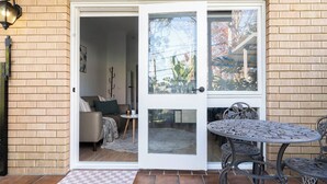 A patio with a table and chairs, a door leading to an interior space with a sofa and a lamp. - Warrina Nook A Cosy Bushland Escape for Two (Chatswood West)