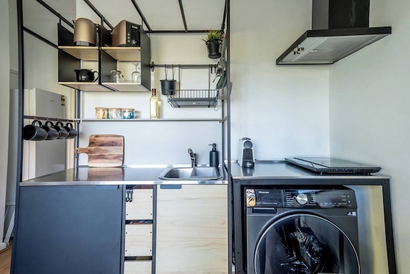 A compact kitchen with a washing machine, a coffee maker, and a laptop on the counter.