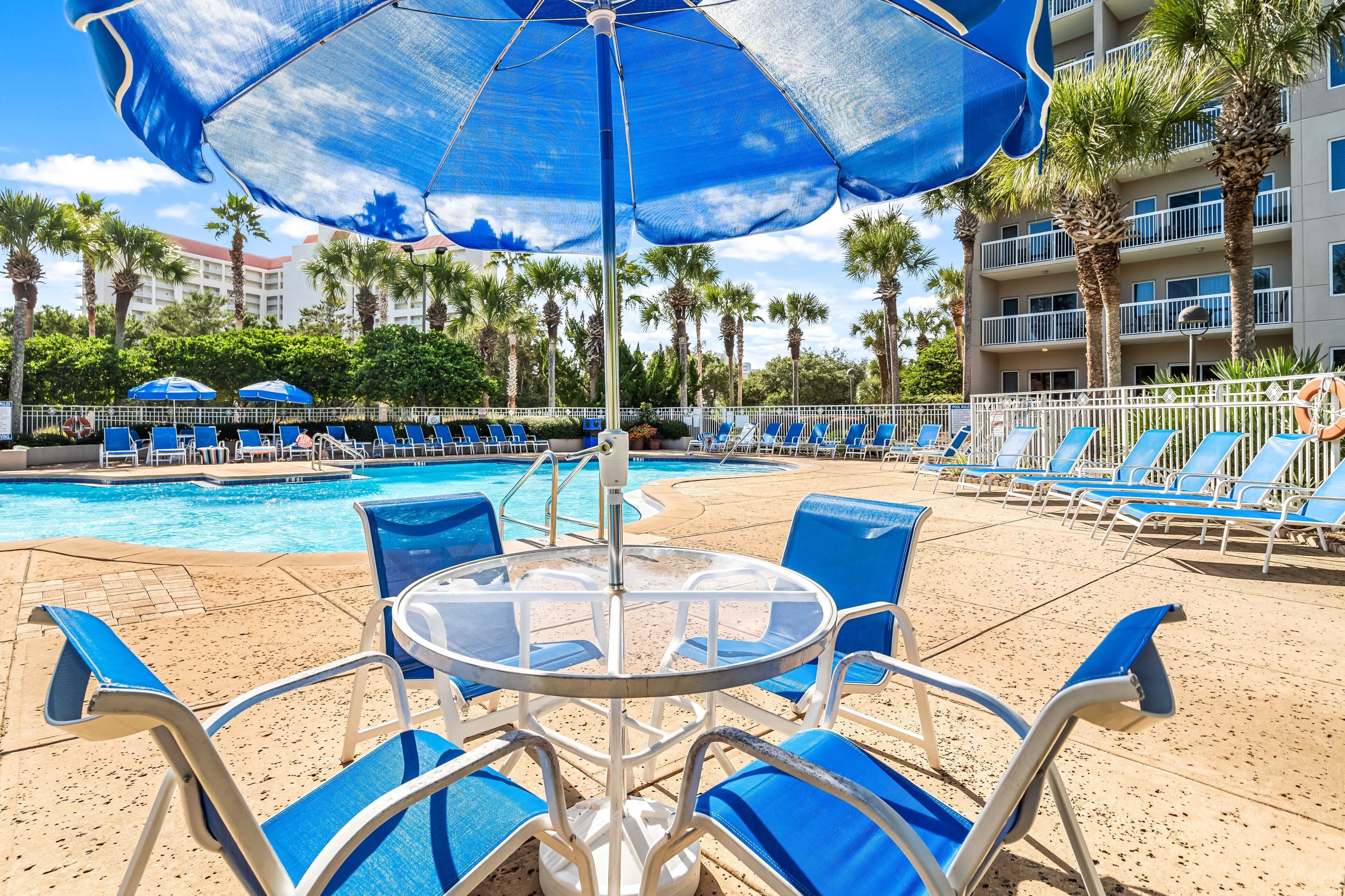 A poolside area with blue chairs and umbrellas, a pool, and a building with balconies.