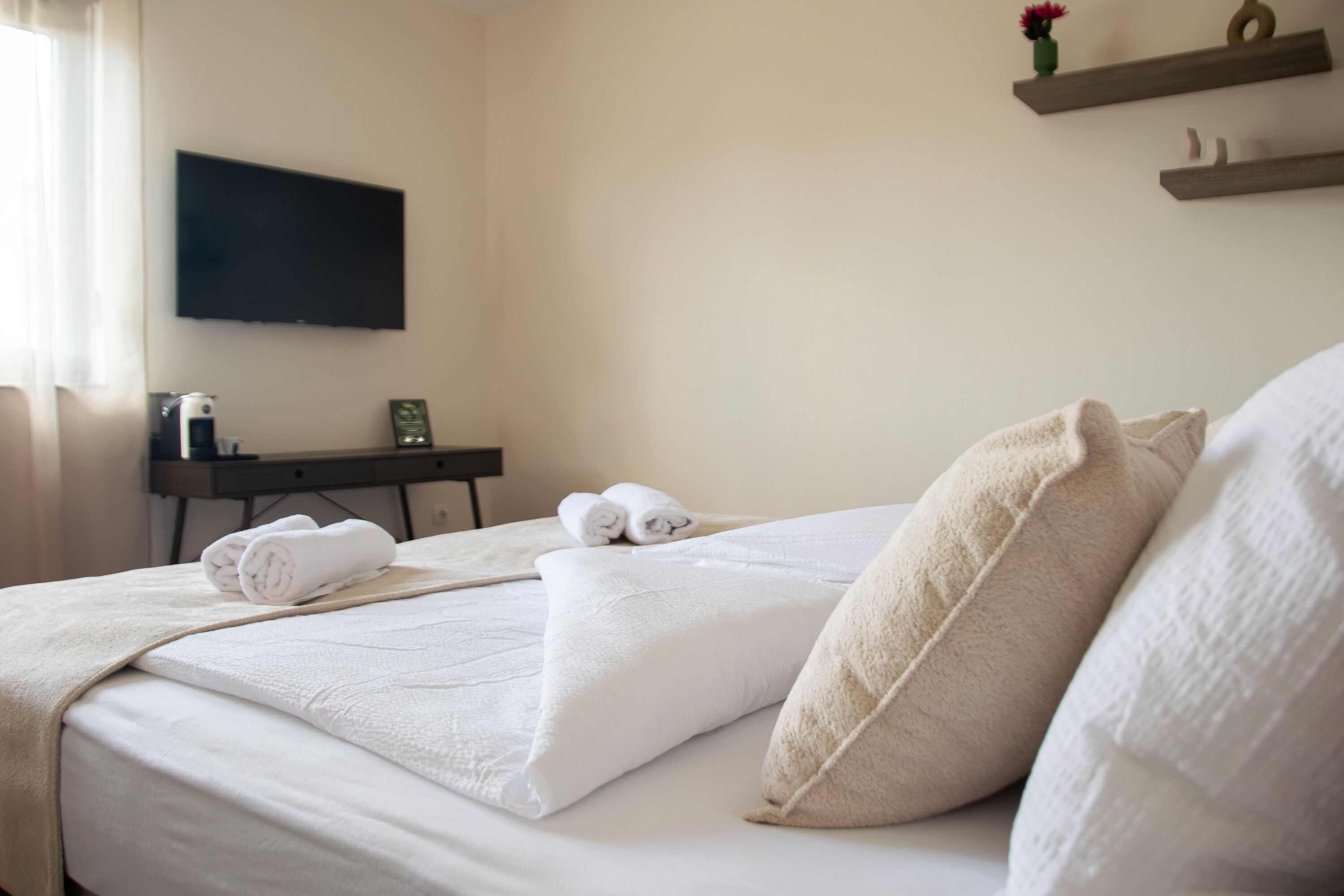Un lit bien fait, avec du linge de lit blanc et des oreillers, un téléviseur à écran plat fixé au mur, et un petit bureau avec une lampe et un tableau encadré.