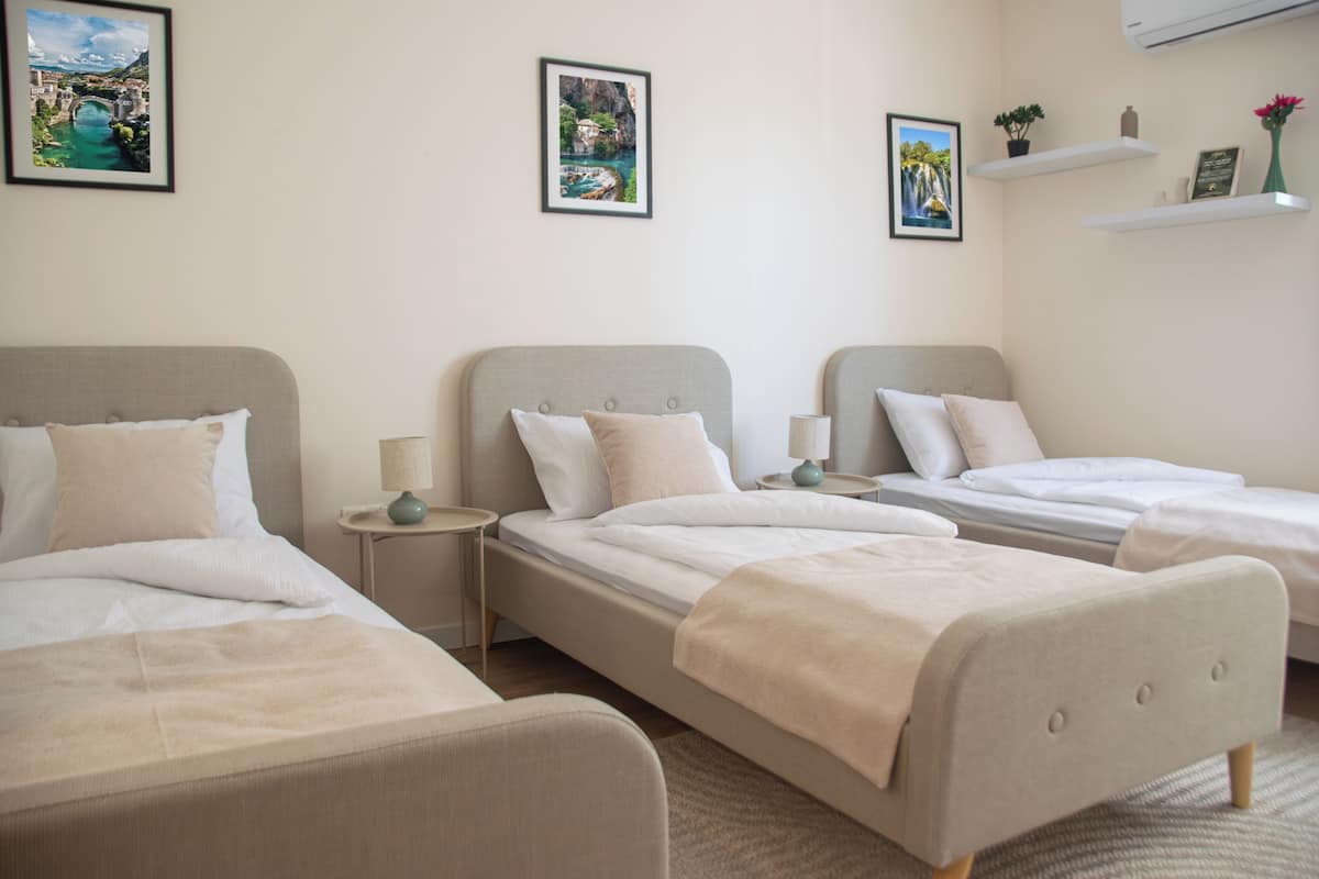 A hotel room with three beds, a small round table, a lamp, and two wall-mounted shelves with decorative items.