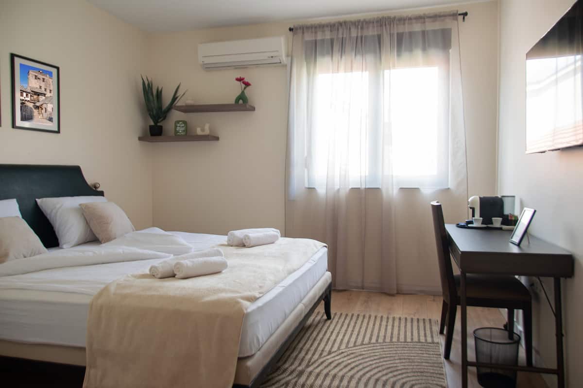 A hotel room with a bed, a desk, a chair, a window with curtains, a potted plant, and a framed picture on the wall.