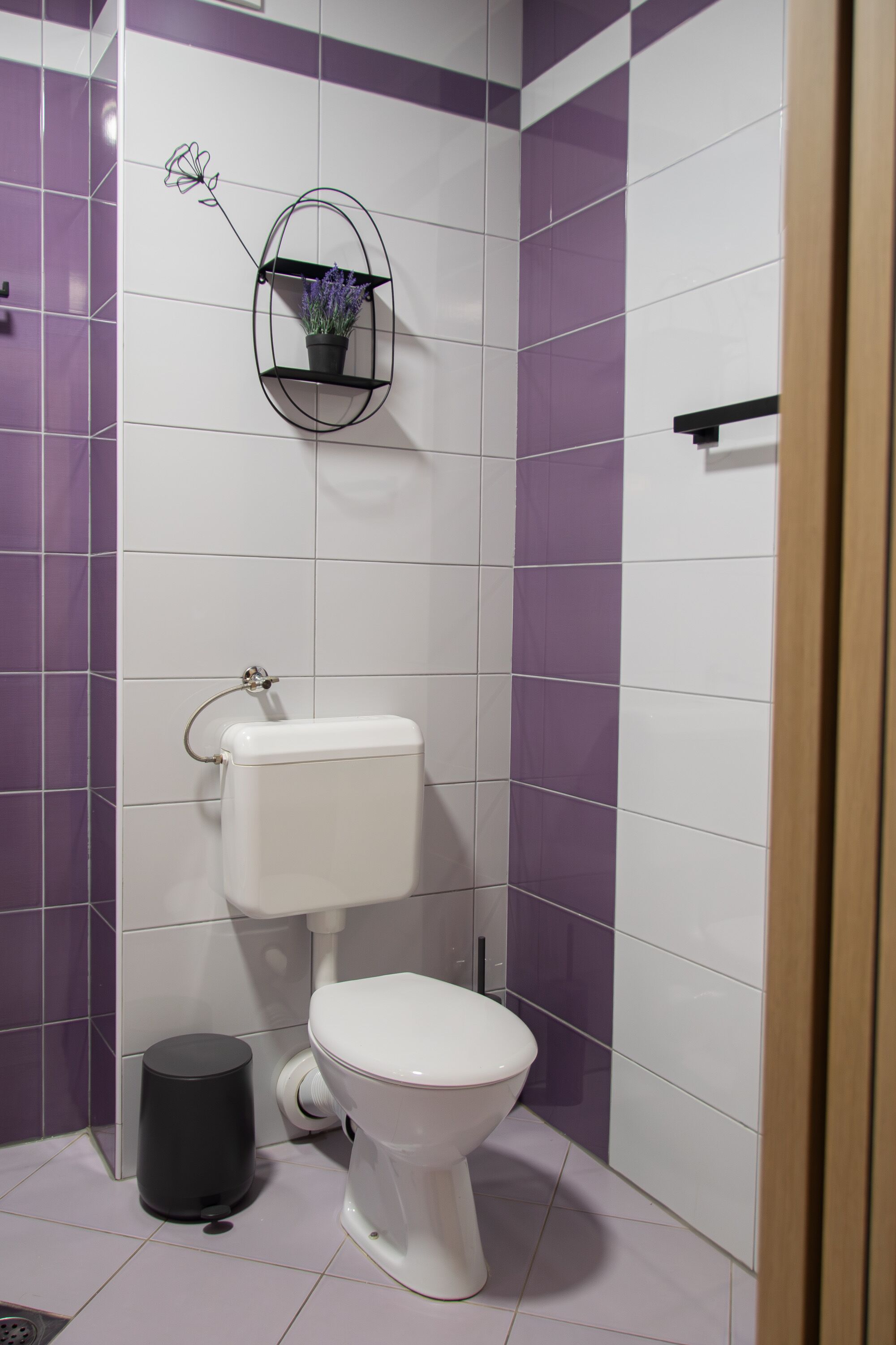 Une salle de bain avec des toilettes, une poubelle noire et une étagère fixée au mur, sur laquelle est posée une fleur.