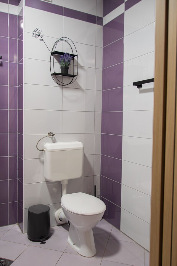 Un baño con inodoro, un bote de basura negro y un estante en la pared con una flor.