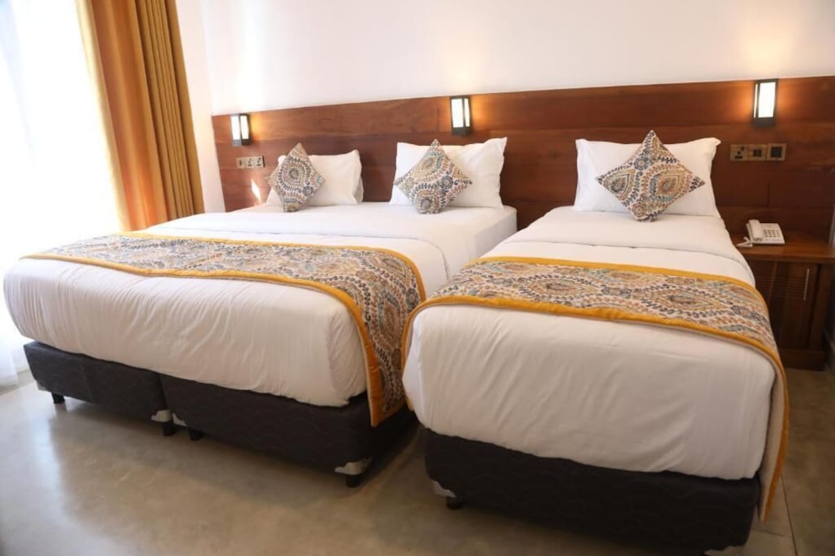 A hotel room with two beds, each with a patterned bedspread and pillows, a wooden headboard, a bedside table with a phone, and a window with curtains.