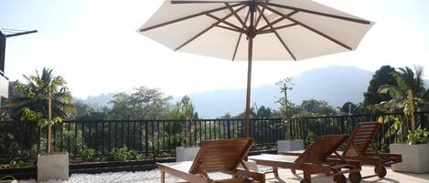 A rooftop terrace with white pebbles, two wooden lounge chairs, a large umbrella, and a view of mountains.