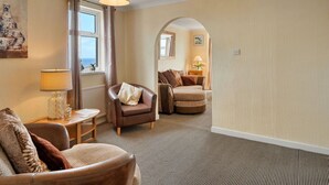 A living room with a sofa, armchair, and a lamp. There is a window with curtains, a painting on the wall, and a carpeted floor. - Lovely apartment for 4 guests with WIFI (St Ives)