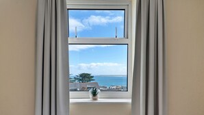 A room with a window overlooking a body of water and a tree, a radiator below the window, and a bed with a white cover. - Lovely apartment for 4 guests with WIFI (St Ives)