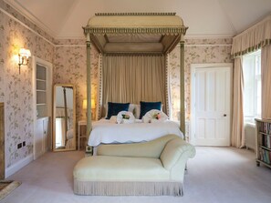 A four-poster bed with a canopy, a sitting area with a patterned ottoman, a full-length mirror, and a bookshelf.