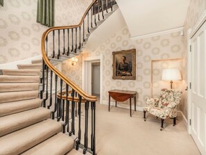 A staircase with a wooden handrail and black balusters, carpeted steps, a small table, a floral chair, a framed portrait, and a lamp.