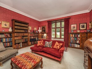 Une bibliothèque avec des étagères, un canapé rouge, un pouf à motifs et une fenêtre avec des rideaux.