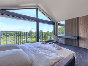 Ein Schlafzimmer mit einem großen Bett, einem Schreibtisch und Blick auf Grün durch große Fenster.