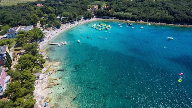 Luftaufnahme einer Küstenregion mit klarem türkisblauem Wasser, einem Sandstrand und einem kleinen Steg.