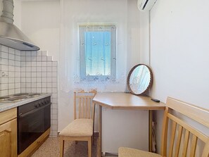 Eine kleine Küche mit Herd, einem Fenster mit Spitzenvorhängen, einem Holzstuhl und einem Spiegel auf einem Tisch.