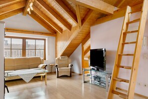 Ein Loft-Zimmer mit Holzbalkendecke, einer Couch, einem Sessel, einem Fernseher auf einem Glastisch und einer Holzleiter.