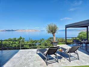 Eine Terrasse mit zwei Liegestühlen, einem Tisch mit Obstschale und einem in einem Blumentopf stehenden Olivenbaum.