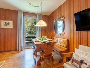 Ein Zimmer mit Holzwänden, einem Esstisch für vier Personen, einer Couch, einem an der Wand montierten Fernseher und einem Fenster mit Blick auf Bäume.