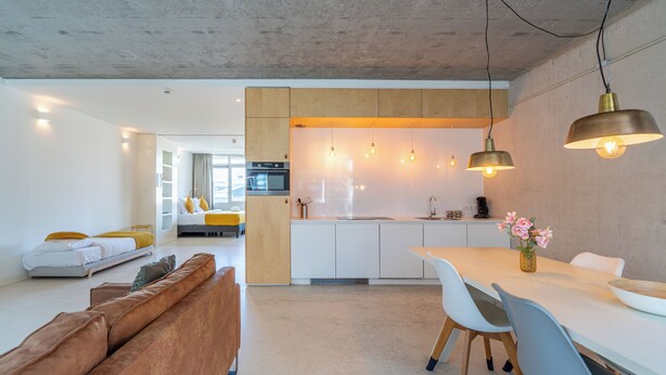 Una cocina moderna con iluminación colgante, una zona de comedor con mesa y sillas blancas, y una sala de estar con un sofá marrón.