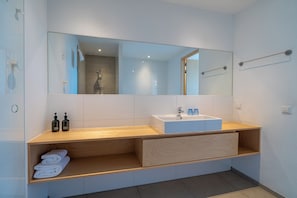 A modern bathroom with a large mirror, a white sink, wooden shelves, and a towel rack.