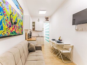 Ein kompakter Wohnbereich mit Sofa, Esstisch, Stühlen, Küchenzeile und einem an der Wand befestigten Fernseher.