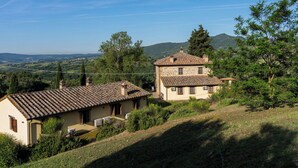 Ein zweigeschossiges Steinhaus mit Ziegeldach, umgeben von viel Grün und Hügeln im Hintergrund.