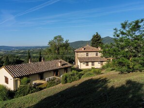 Ein zweigeschossiges Steinhaus mit Ziegeldach, umgeben von viel Grün und Hügeln im Hintergrund.