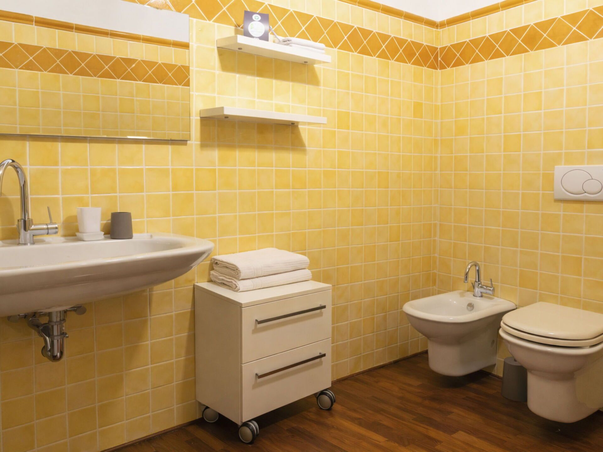 A bathroom with yellow tiles, a sink, a bidet, a toilet, and a storage cart.