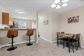 A kitchen with wooden cabinets, a microwave, and a refrigerator. There are two bar stools and a dining table with chairs.