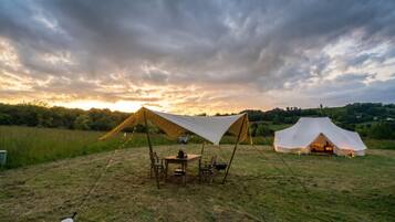 A tented camping area with a table and chairs under a canopy, surrounded by grassy fields and a cloudy sky.