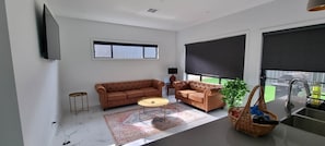 A living room with two sofas, a coffee table, a TV, a window with blinds, a plant, and a kitchen area with a sink.