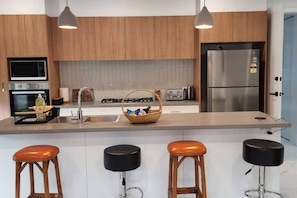 A modern kitchen with wooden cabinets, a stainless steel refrigerator, a microwave, and a sink. There are three stools and a basket on the counter.