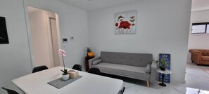 A modern living room with a grey sofa, a dining table with a potted plant, a wall-mounted artwork, and a small table with a sign.