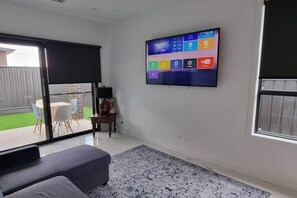 A modern living room with a flat-screen TV mounted on the wall, a sofa, a patterned rug, and a sliding glass door leading to a patio with outdoor furniture.