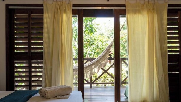 A room with a bed, a hammock, and a view of a wooden deck and greenery.